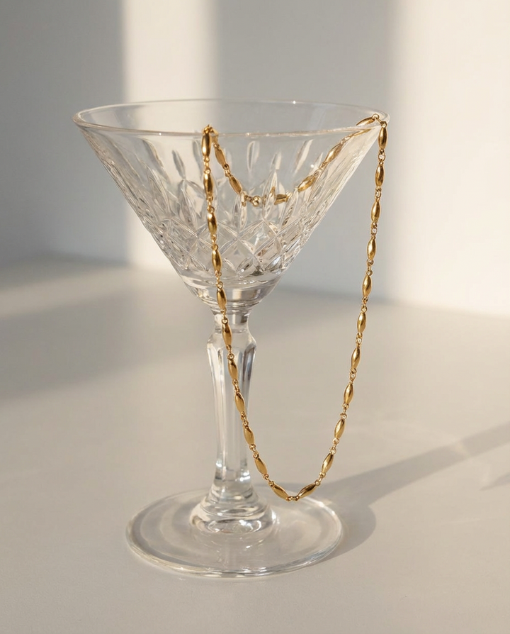 The gold oval link chain necklace draped over a glass, styled in a clean and elegant decorative setup.