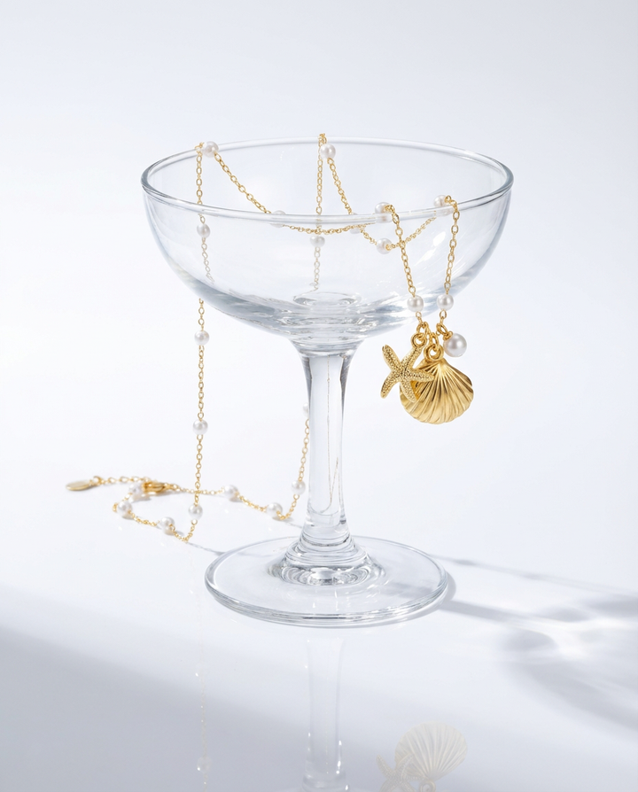 The gold seashell and starfish pendant necklace draped over a glass, styled in an elegant decorative setting.